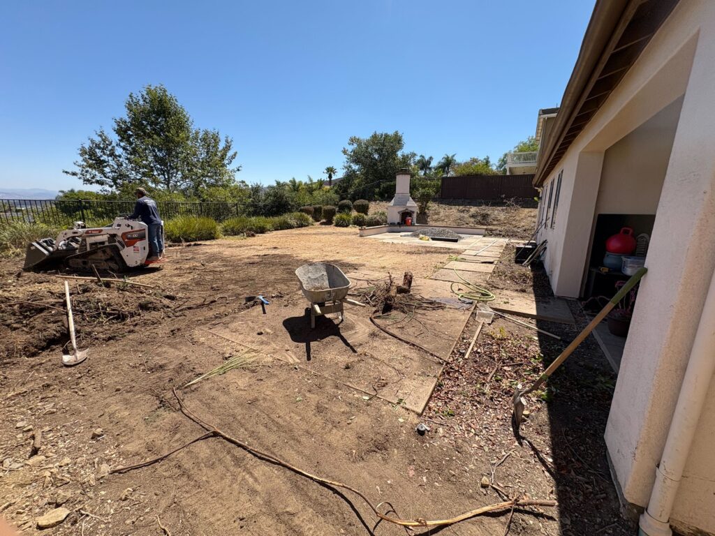 Backyard construction site with turf/topsoil removed and equipment staged for site work. An existing outdoor fireplace...