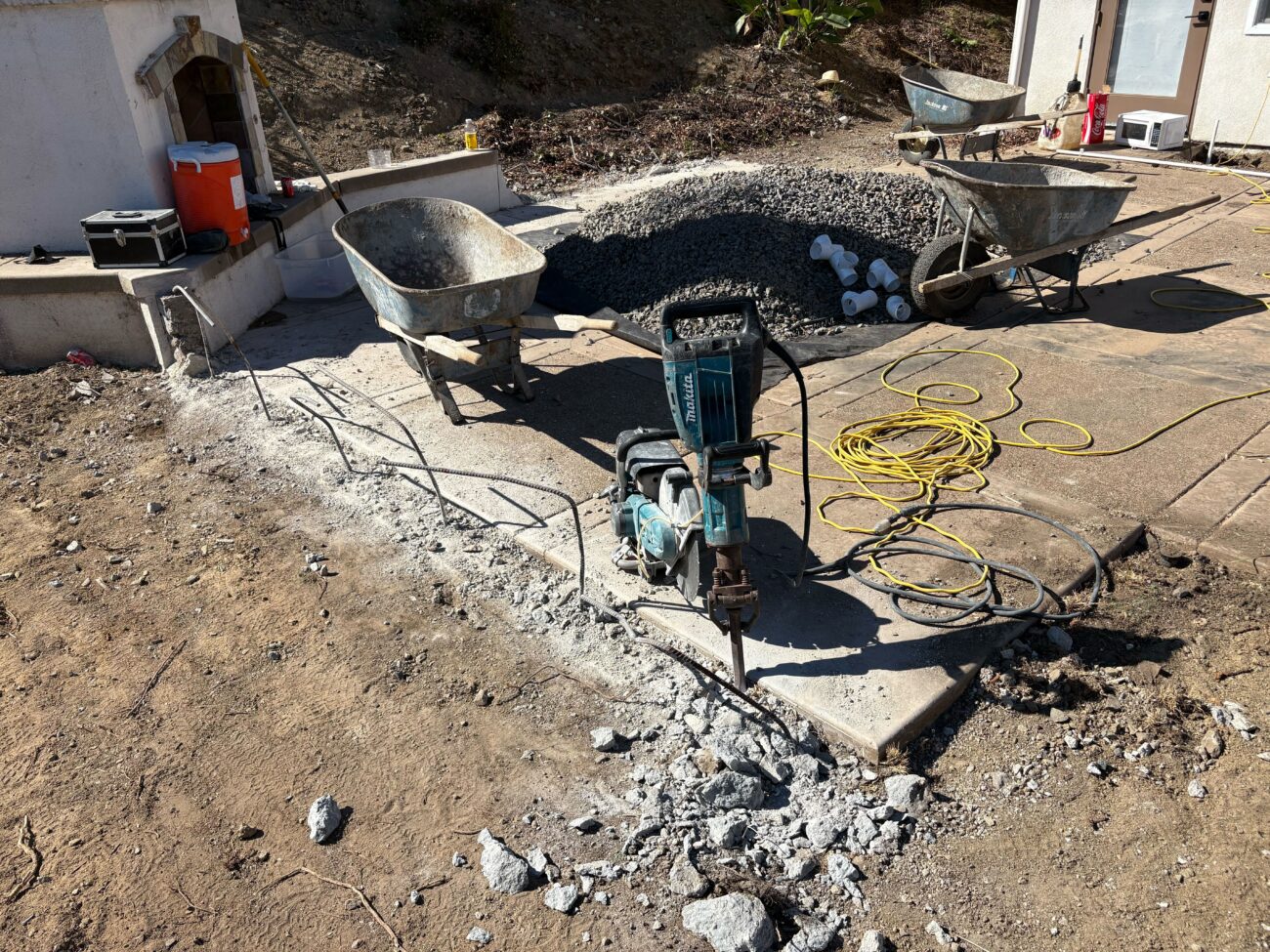 Backyard patio concrete is actively being demolished with a handheld breaker/jackhammer and scattered concrete rubble....