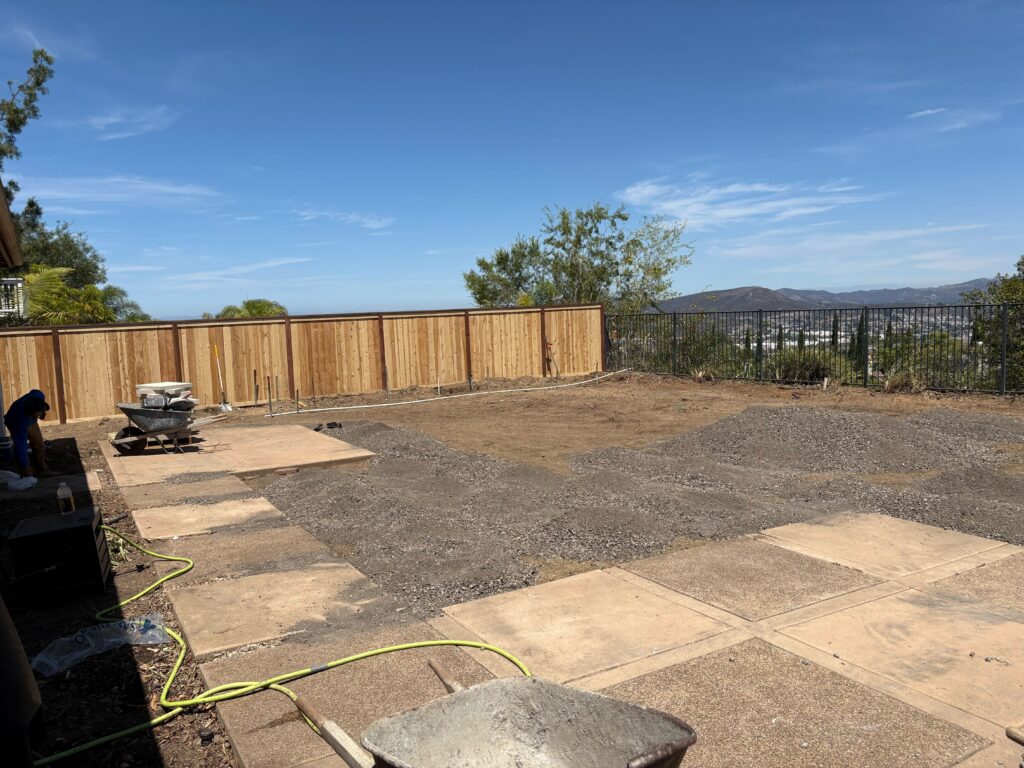 Residential backyard under construction showing a compacted gravel base with large concrete paver pads laid out across...