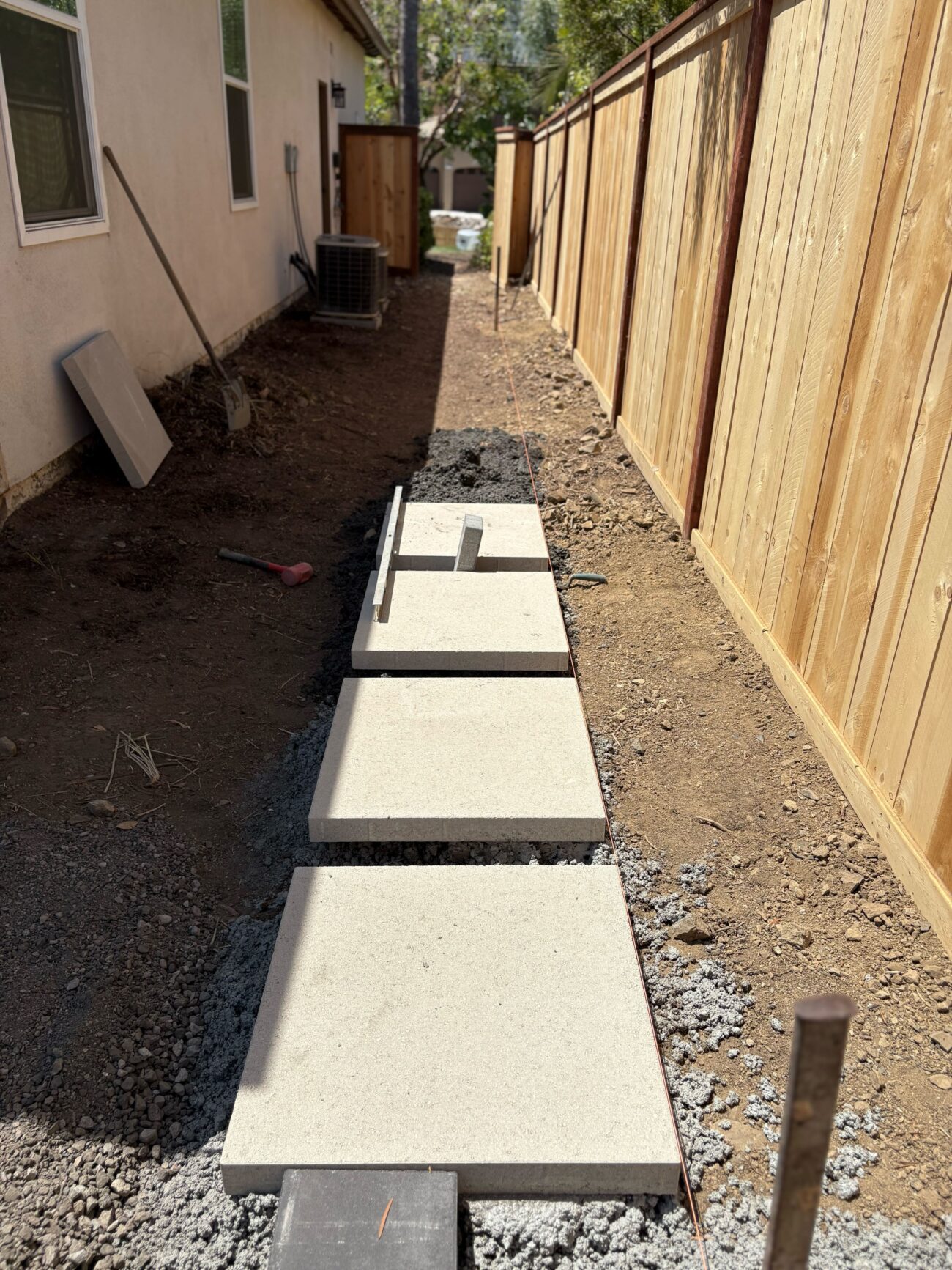 Narrow residential side yard with large concrete pavers laid in a single-file walkway along a wooden fence. The pavers...