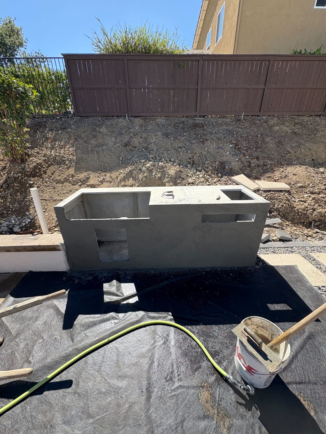 Partially constructed outdoor kitchen/island built from concrete block with a stucco finish, showing openings for a...
