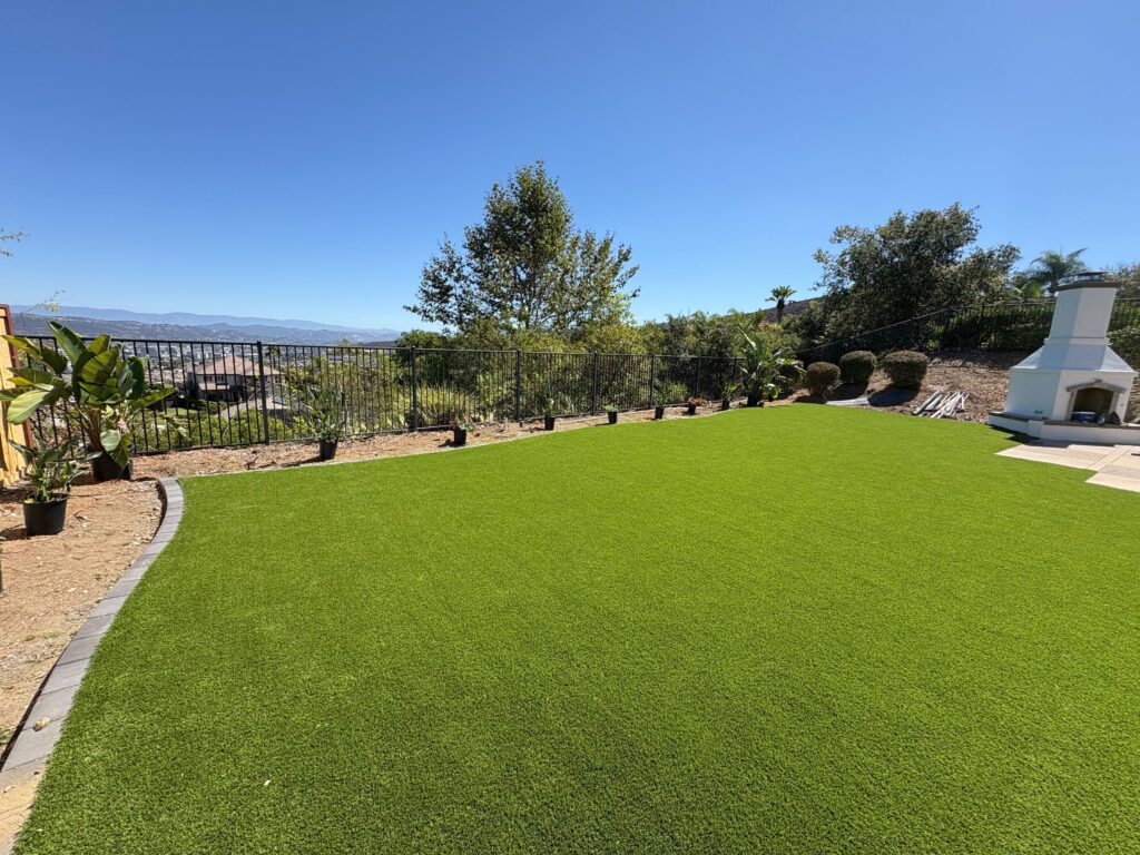 Residential backyard with newly installed artificial turf framed by a paver edge and adjacent concrete/paver patio with...