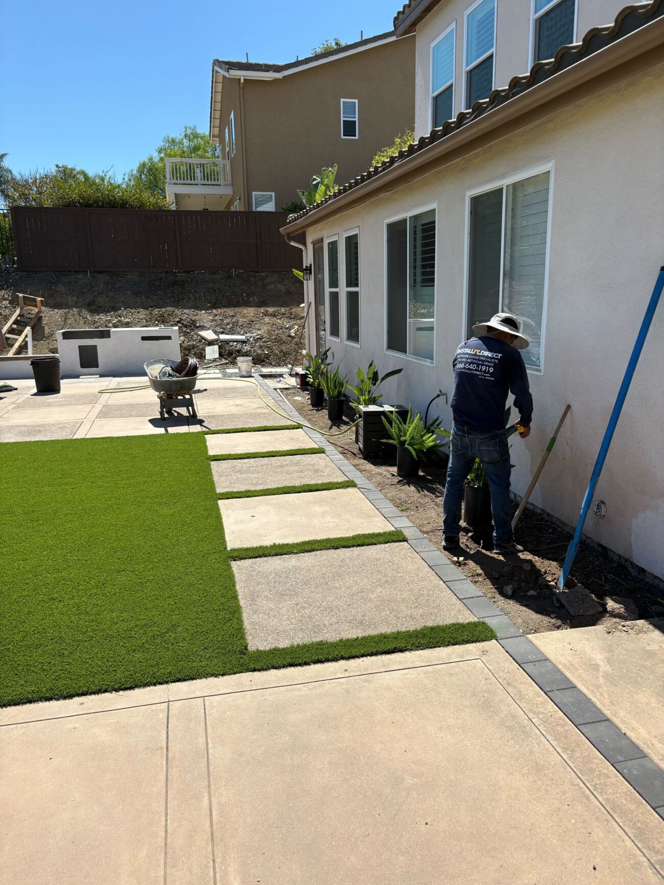 Backyard scene showing artificial turf installed with rectangular concrete stepping pads and a paver border adjacent to...