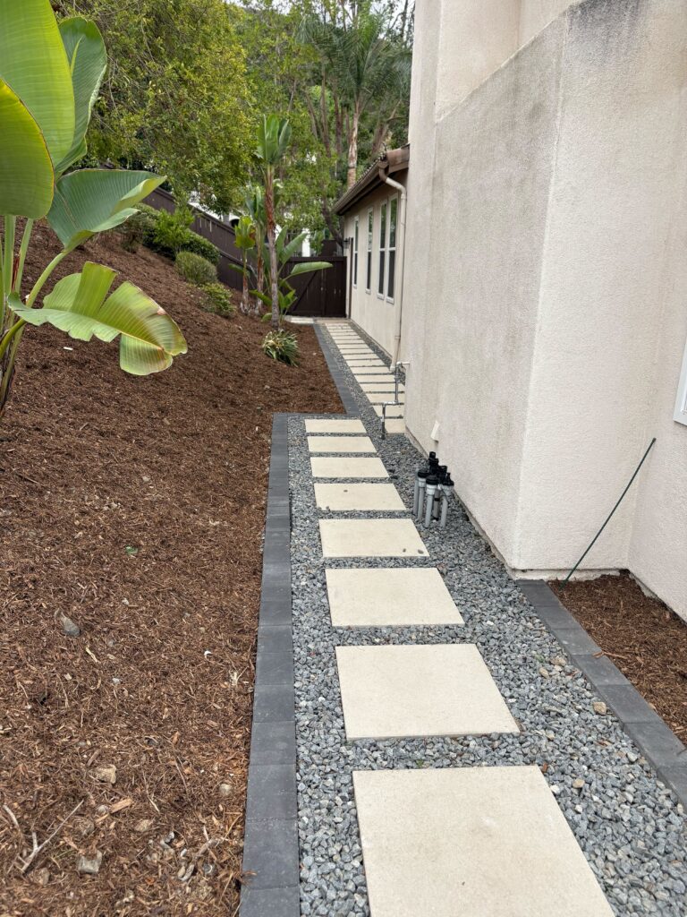 Finished side-yard walkway featuring spaced concrete stepping pads set in decorative gray gravel with a dark...
