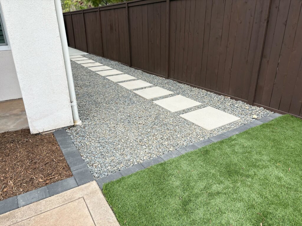 Finished side-yard installation showing rectangular concrete stepping pads set in compacted decorative gravel with a dark...