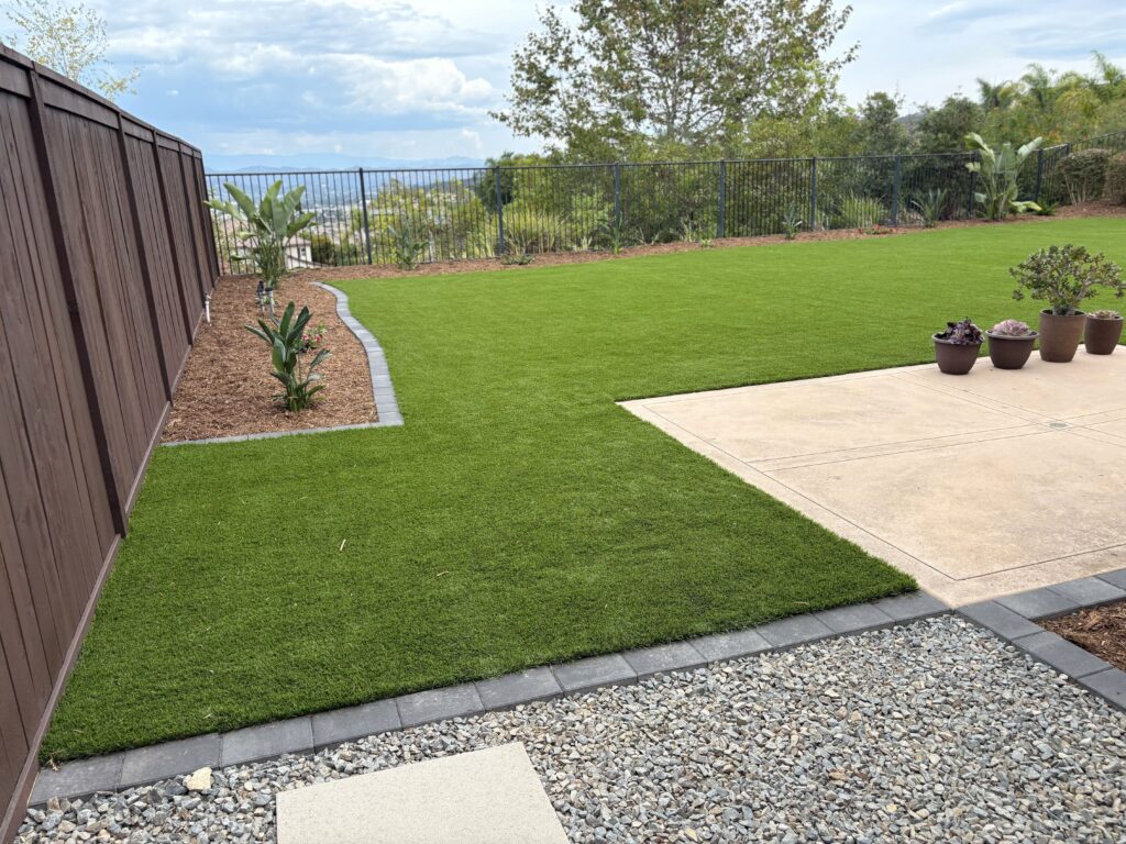 Finished backyard landscape showing newly installed artificial turf edged with a paver border and an adjacent concrete...