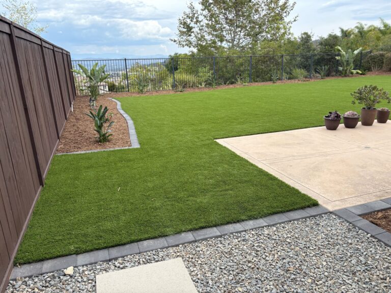 Finished backyard landscape showing newly installed artificial turf edged with a paver border and an adjacent concrete...