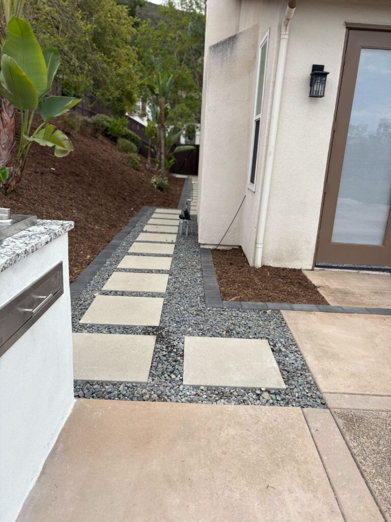 Concrete paver stepping stones set in decorative gravel form a linear walkway along the side of a residence, bordered by...