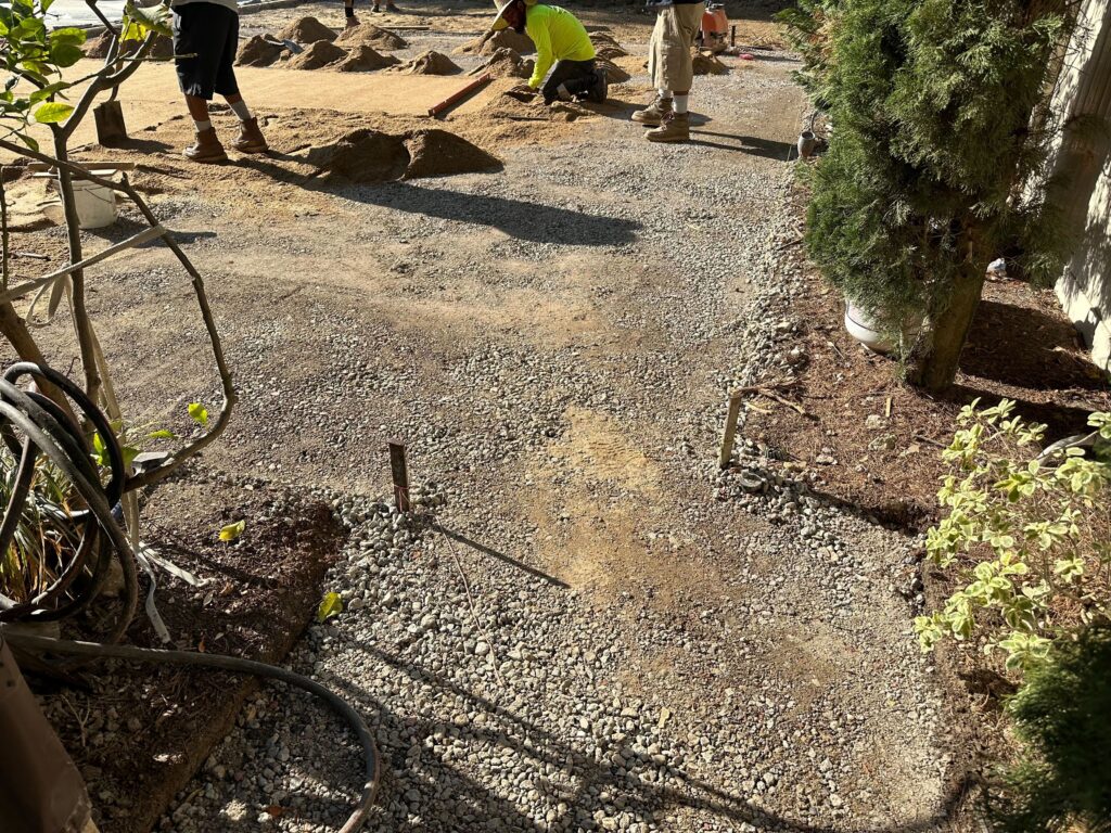 Crew members are spreading and screeding compacted aggregate and bedding material in a backyard area to prepare the...