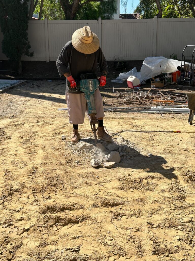 Worker operating a jackhammer to break up a concrete slab in a residential backyard. Exposed sandy subgrade and scattered...