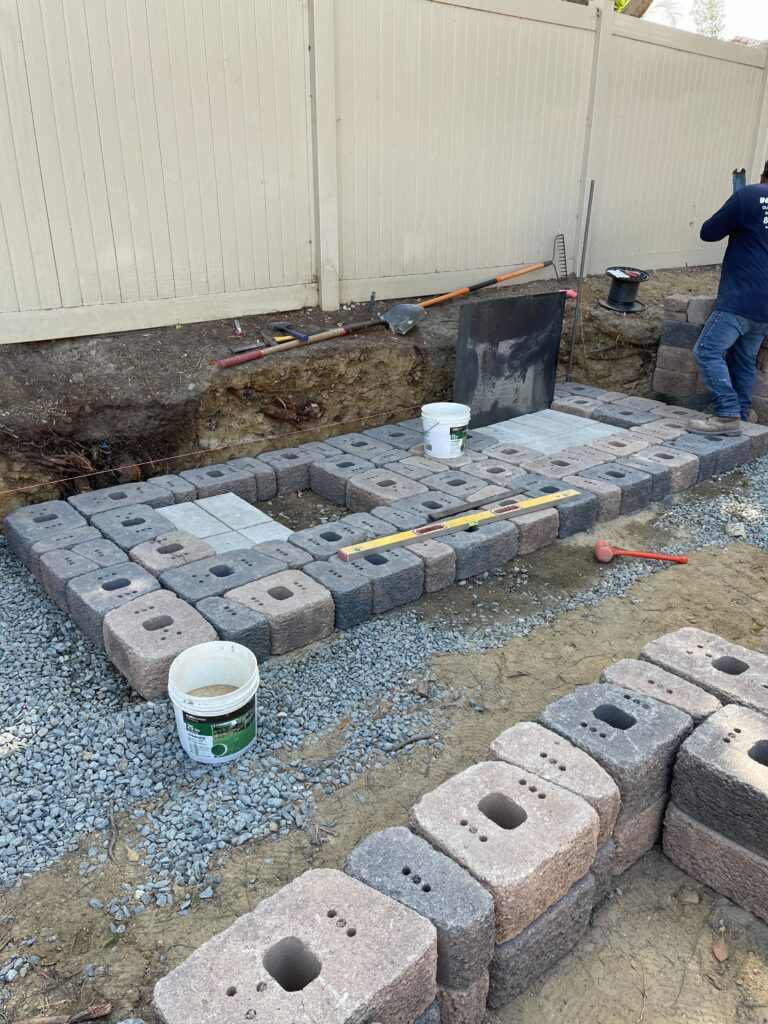 Segmental retaining wall block installation in a residential side yard; hollow concrete wall units are being set on a...