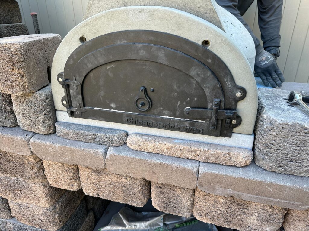 Close-up of a wood-fired pizza oven insert being built into a segmental block surround. Installer hands and tools are...