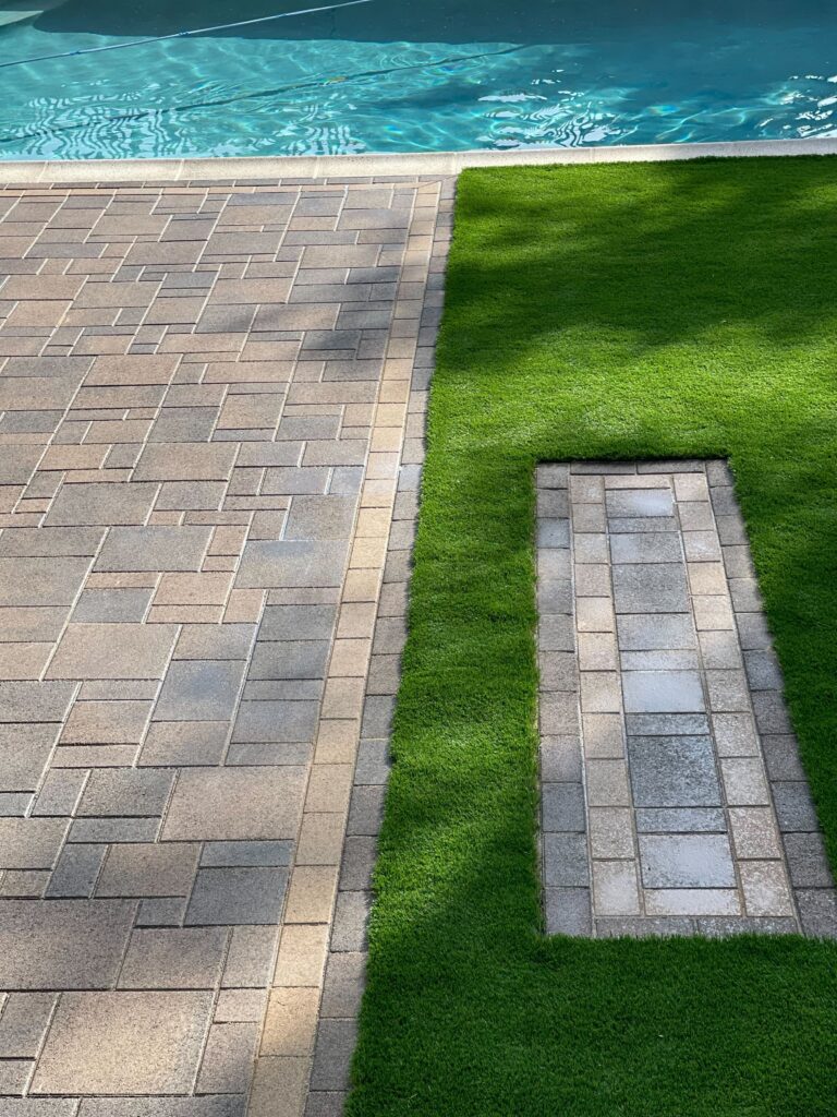Completed outdoor installation showing an interlocking paver patio abutting artificial turf with a pool deck visible at...