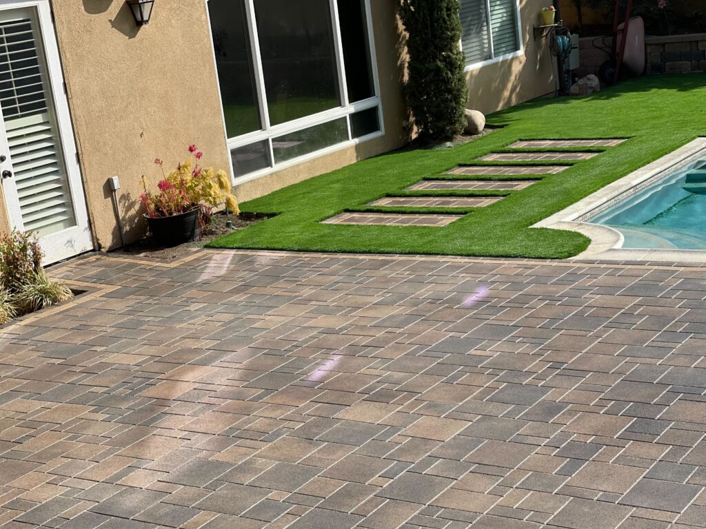 Finished interlocking paver patio adjacent to a residential pool with artificial turf and integrated paver stepping...