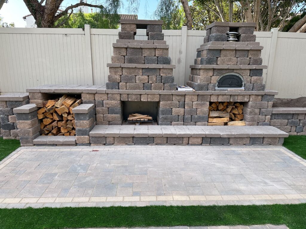 Completed masonry outdoor kitchen and fireplace assembly built from segmental concrete pavers with integrated wood...