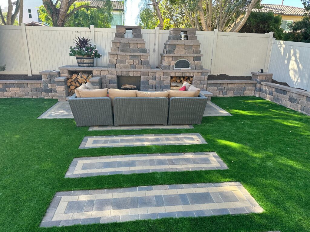Backyard installation showing synthetic turf with rectangular interlocking paver stepping pads leading to a segmental...