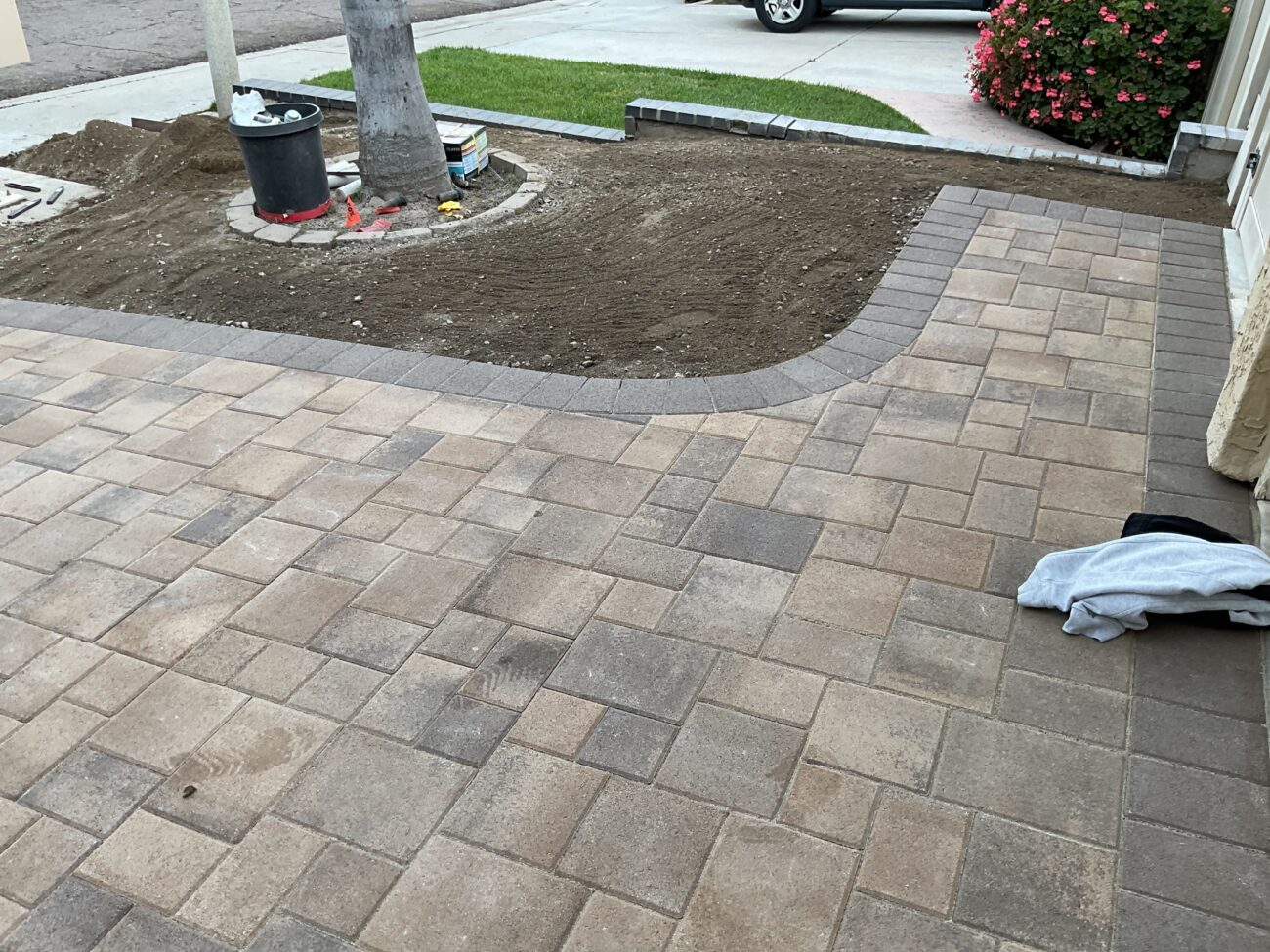 Interlocking paver patio with a darker soldier-course border installed adjacent to a concrete driveway and lawn strip. A...