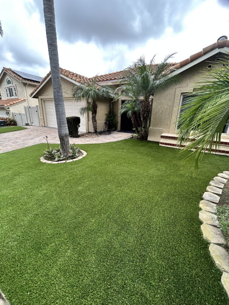 Front yard of a residential property showing a completed artificial turf installation with stone edging and an...