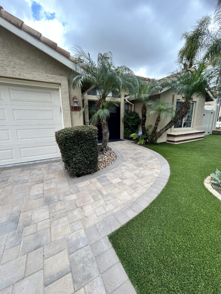 Completed front-entry landscape showing interlocking pavers forming a curved driveway/walkway and adjacent artificial...