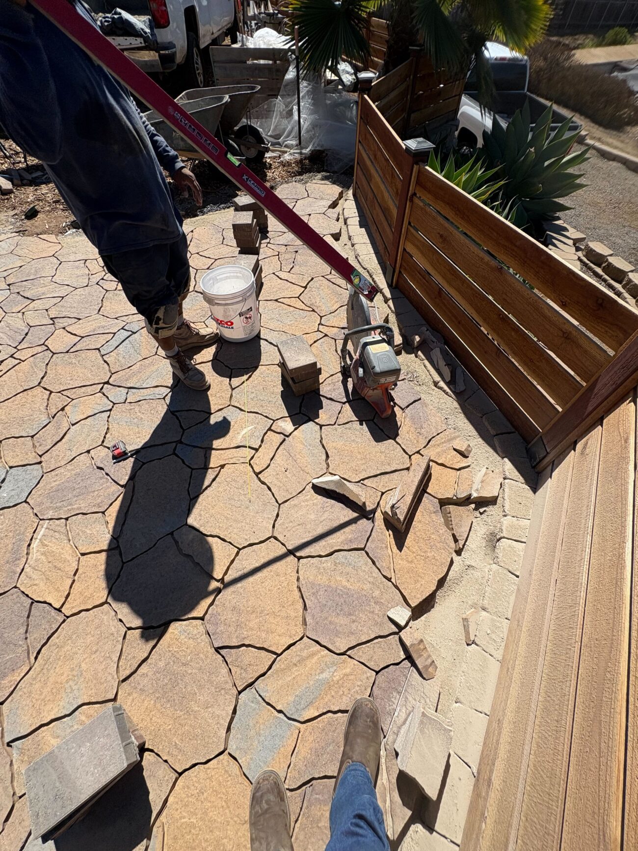 Crew is actively installing irregular flagstone pavers for a residential patio, with a handheld saw/grinder and bucket visible while stones are being cut and fit at the edge next to a wooden fence. El Cajon project by Install It Direct.