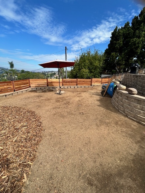Backyard construction site showing a compacted dirt subgrade with a completed segmental retaining wall and new horizontal wood fence along the rear. El Cajon project by Install It Direct.