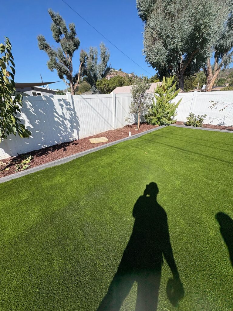 Backyard view of a newly installed artificial turf lawn with clean edge restraint and a mulch planting bed along a white...