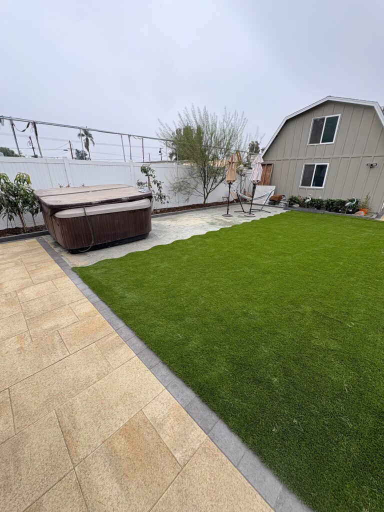 Backyard showing newly installed artificial turf abutting an interlocking paver patio and walkway, with a hot tub...