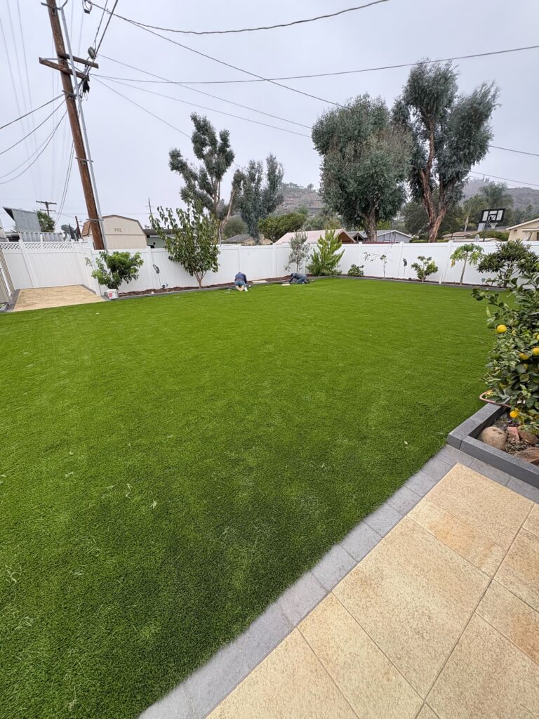 Residential backyard showing a newly installed artificial turf lawn with a paver border and adjacent tiled patio....