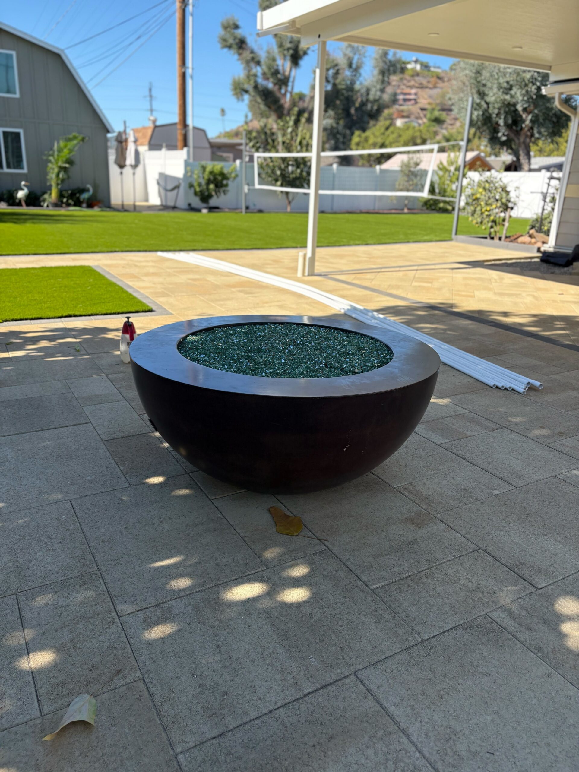 Completed paver patio and adjacent artificial turf in a residential backyard, featuring a large bowl-style fire pit...