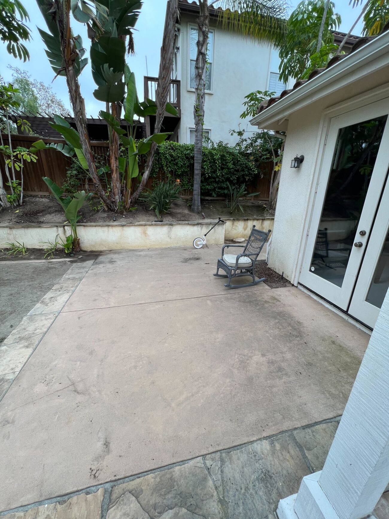 Existing concrete patio slab with visible staining and a stone trim, adjacent to a stucco house and French doors. A low...
