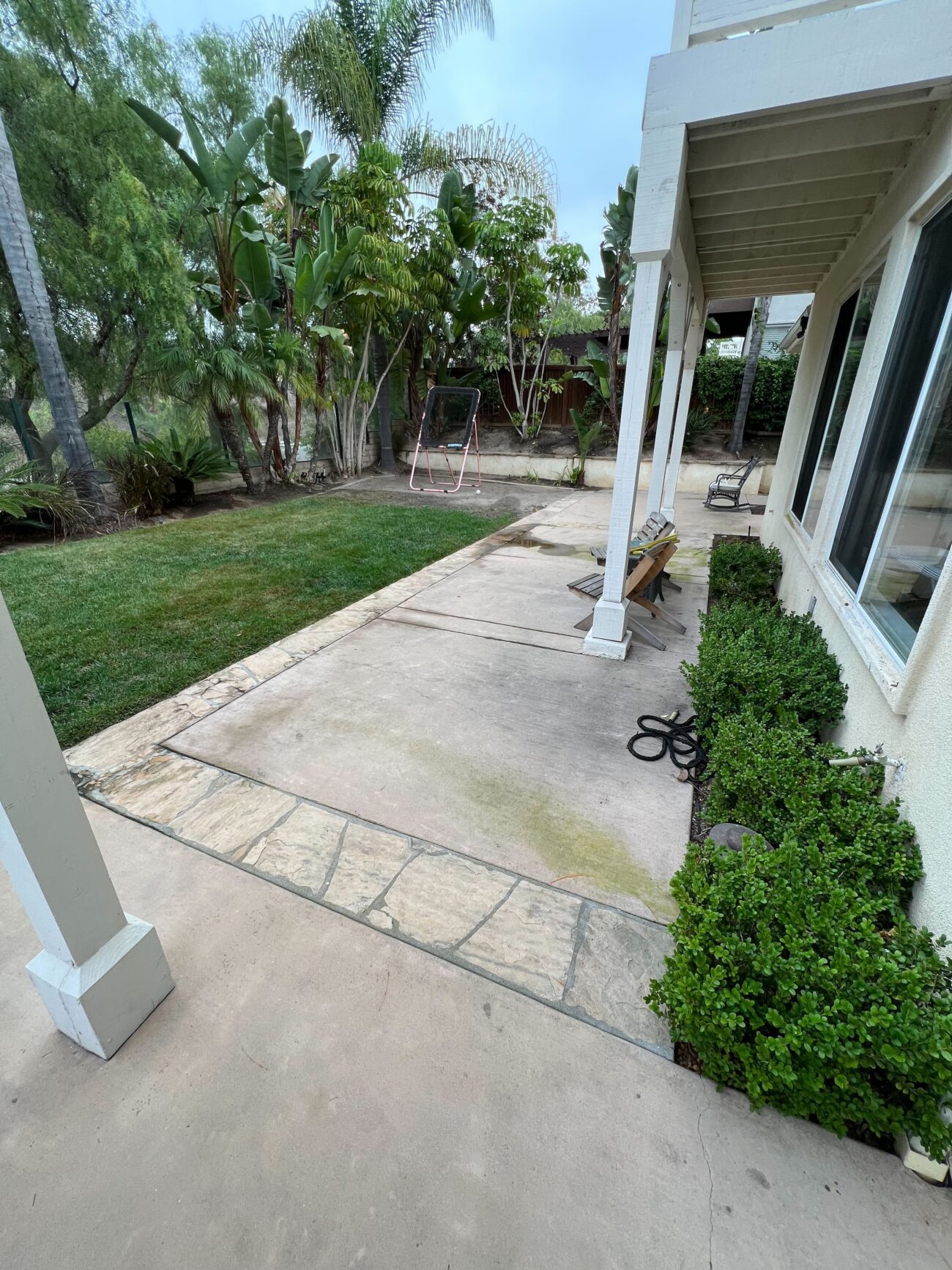 Backyard concrete patio and walkway adjacent to a natural grass lawn and planted shrub border, with a covered porch...