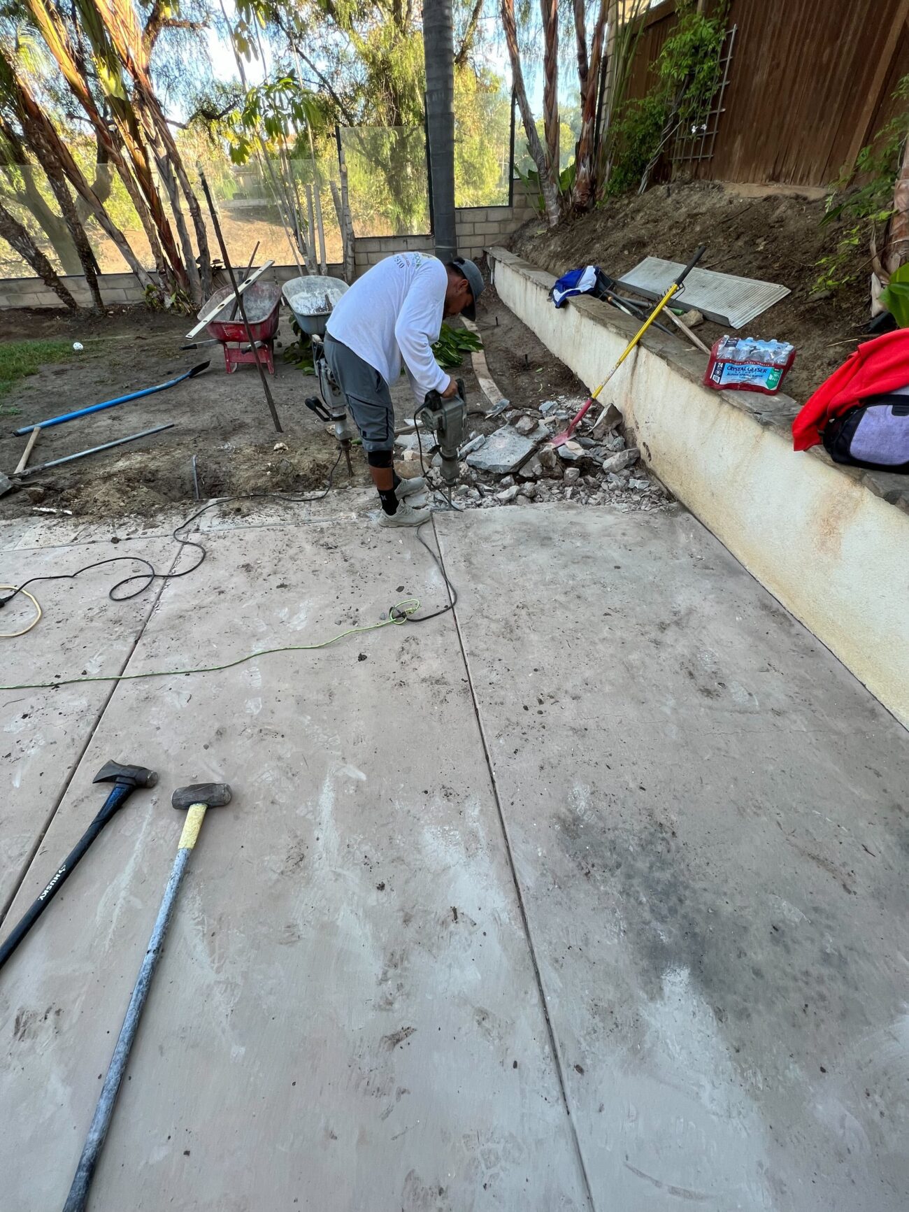 A crew member is jackhammering and breaking out an existing concrete patio slab along a stucco retaining wall in a...
