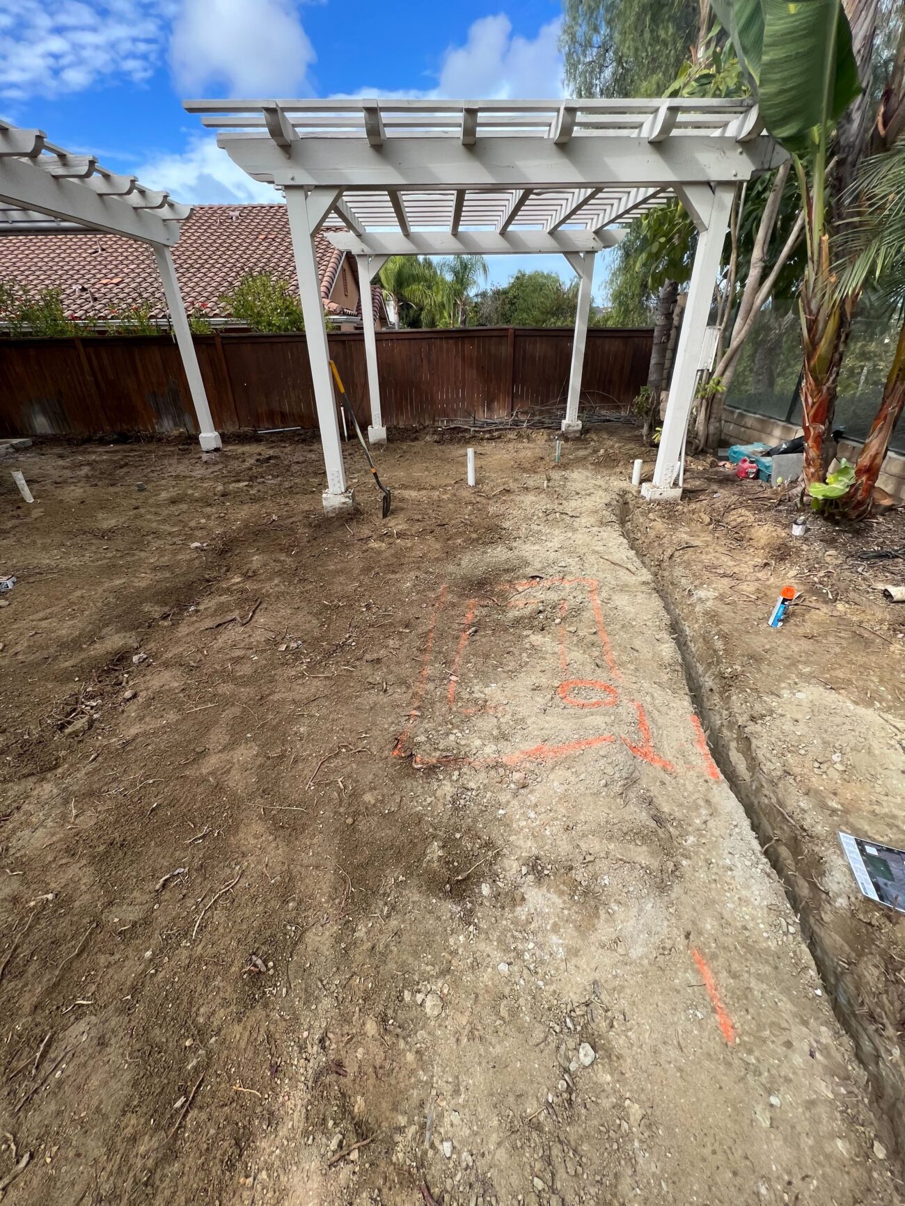 Backyard area cleared to subgrade beneath an existing pergola with a narrow trench excavated and PVC plumbing/utility...