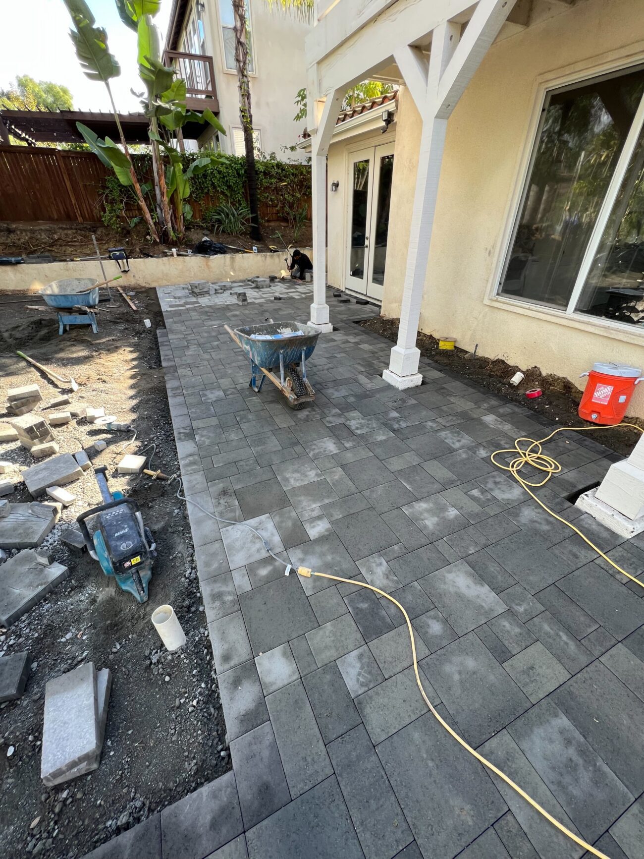 Crew is installing a multi-size modular paver patio adjacent to a house covered porch; dark gray interlocking pavers are...