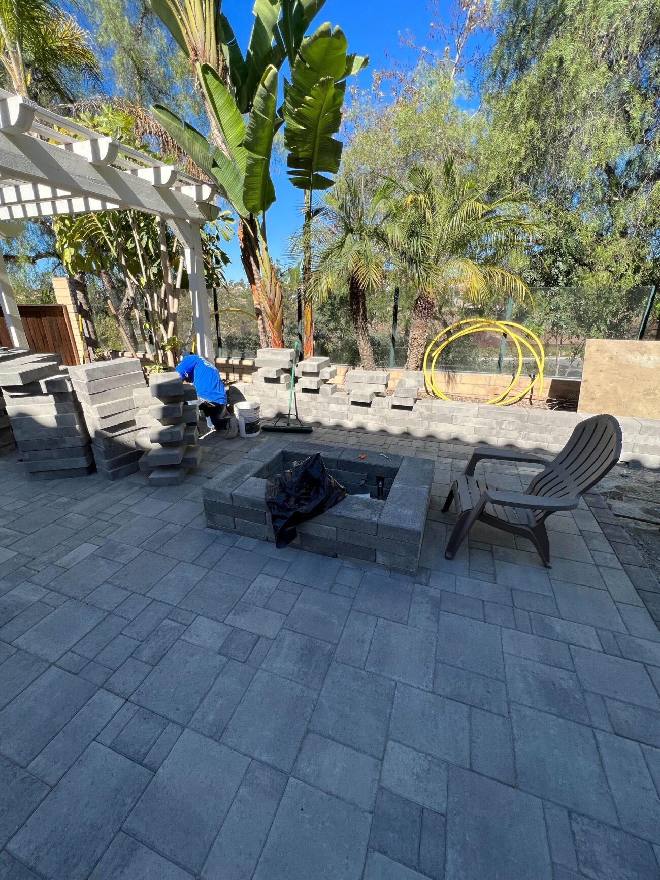 Backyard paver patio under active installation with interlocking pavers laid in a modular pattern. A segmental block fire...