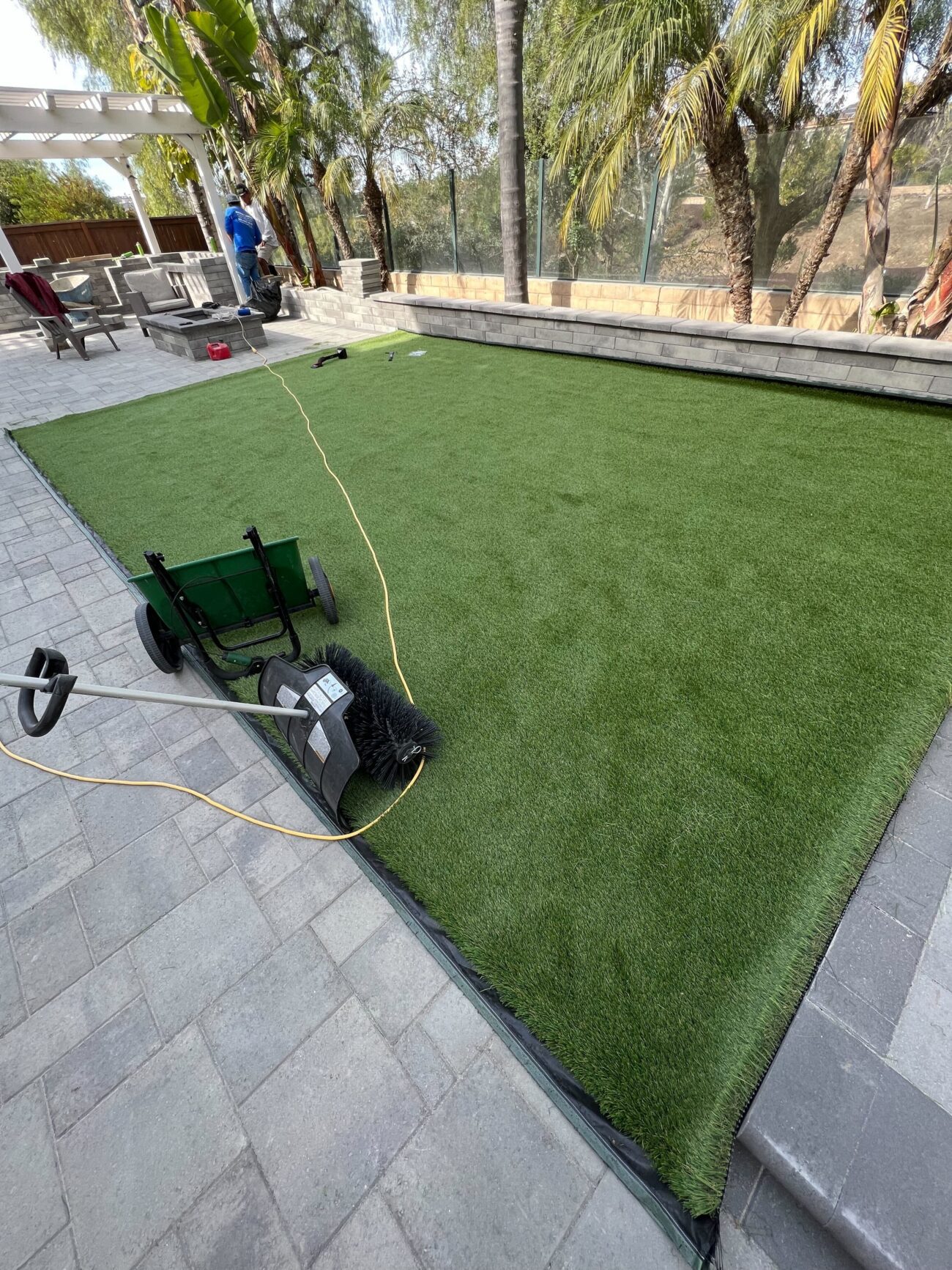 Backyard scene showing freshly laid artificial turf bounded by an interlocking paver perimeter and a low retaining/seat...