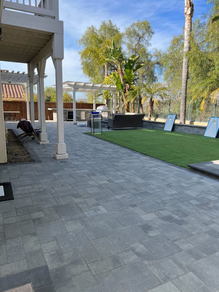 Completed backyard showing a large interlocking paver patio adjacent to a rectangular artificial turf lawn. The space...