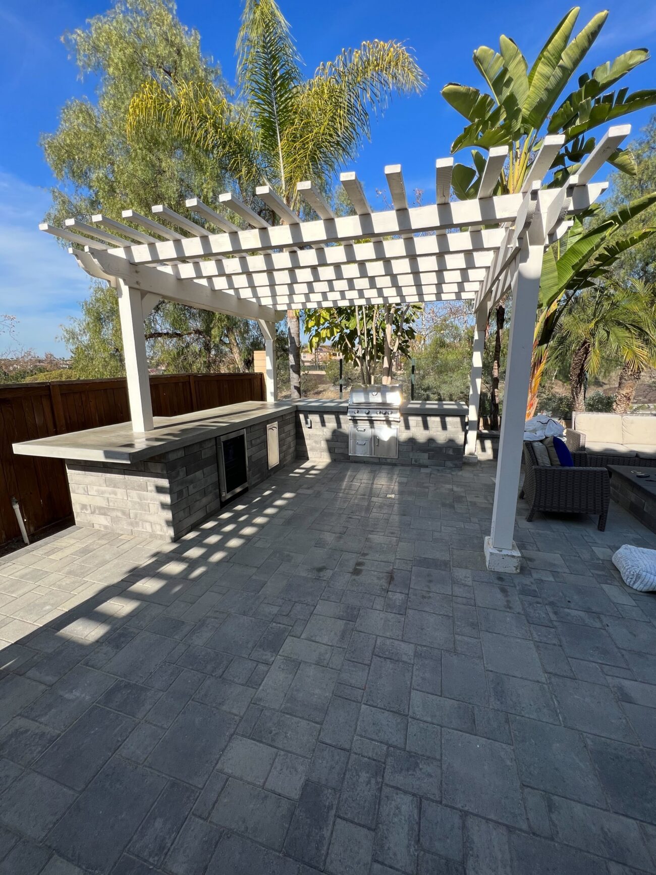 Backyard paver patio with interlocking gray pavers beneath a white pergola, showing a built-in outdoor kitchen island...