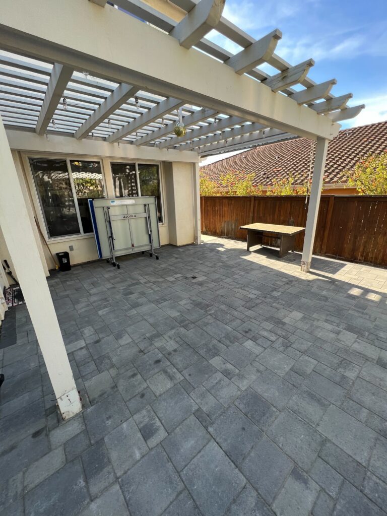 Finished backyard patio showing interlocking concrete pavers installed under a pergola attached to the house. The...