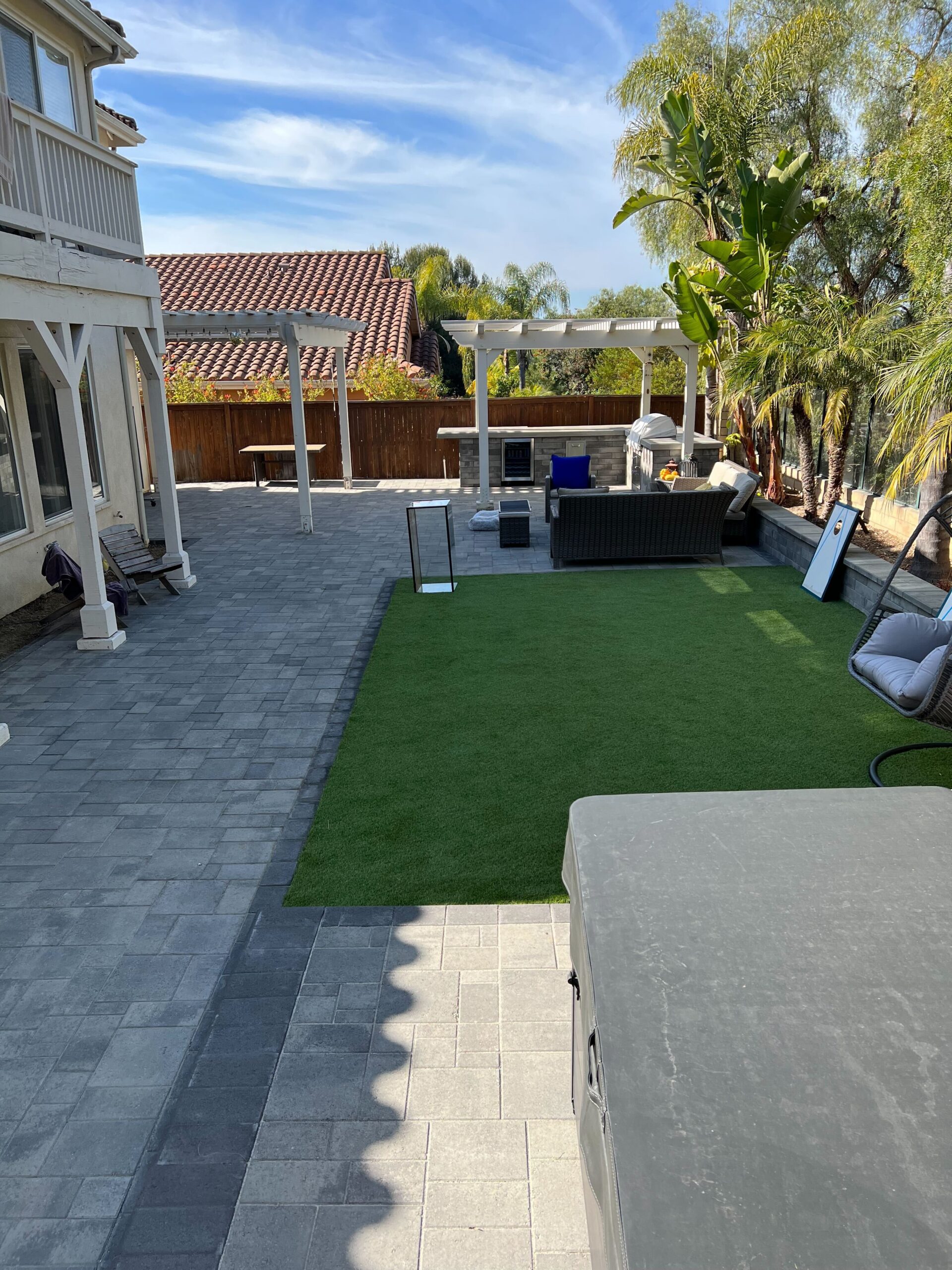 Backyard shows a completed interlocking paver patio with an inset artificial turf area, pergola-covered seating and a...