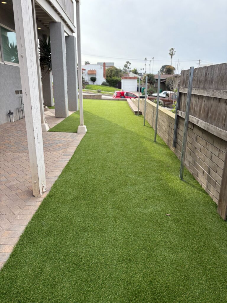 Newly installed artificial turf in a narrow side yard adjacent to an interlocking paver walkway and patio under a raised...