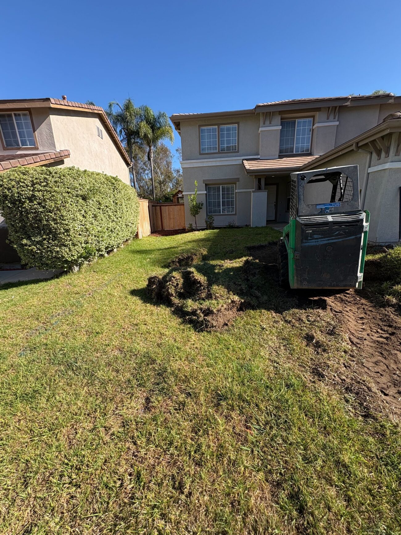 Front yard of a two-story residence where a small skid steer has excavated and removed existing sod, leaving rolled turf clumps and disturbed topsoil. San Marcos project by Install It Direct.