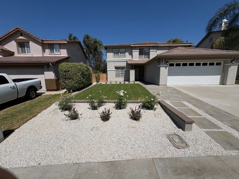 Front yard landscape showing a completed artificial turf lawn flanked by decorative white rock groundcover and a paver edge/walkway adjacent to a concrete driveway. San Marcos project by Install It Direct.
