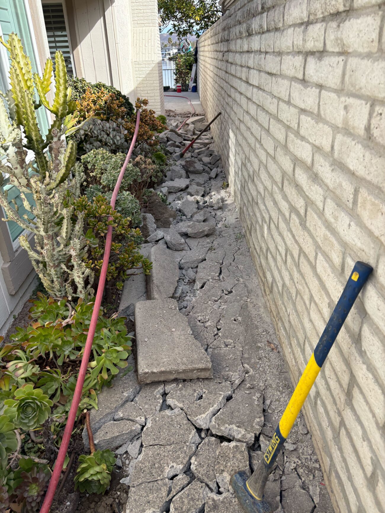 Demolition of an existing concrete walkway in a narrow side yard, showing broken concrete slabs and rubble along the...