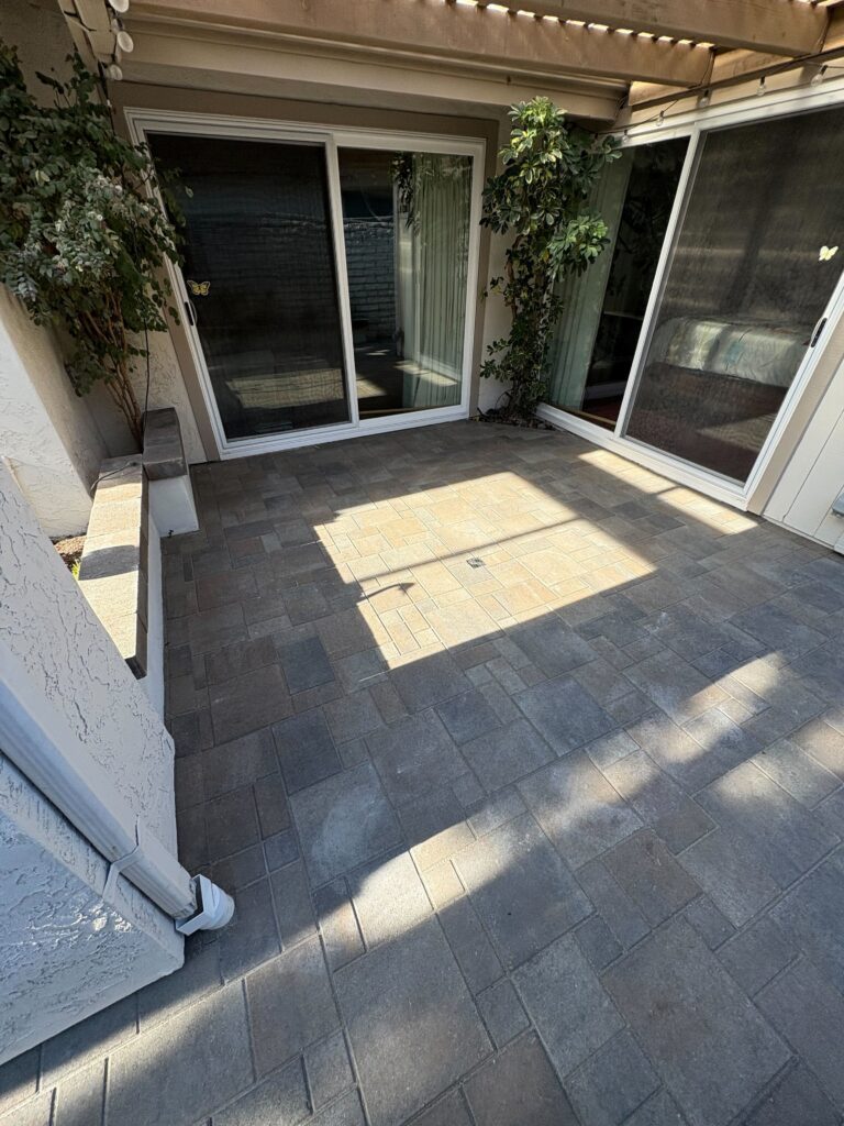 Finished interlocking paver patio under a pergola adjacent to sliding glass doors. The installation includes a central...