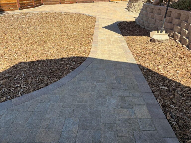 Curved interlocking paver walkway with modular rectangular pavers leads through a backyard landscape with mulch beds and...