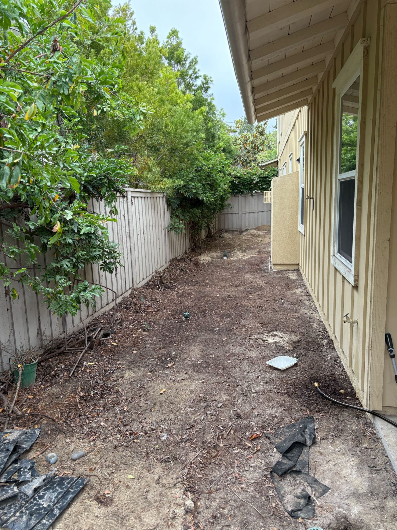 Narrow residential side yard cleared down to exposed soil with remaining landscape debris and torn fabric. The house...