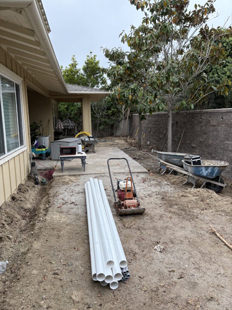 Backyard construction site showing an existing concrete patio and an excavated dirt path adjacent to a segmental...