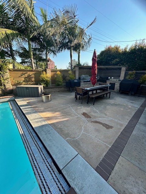 Backyard pool area showing a concrete pool deck and adjacent patio with a brick strip border and outdoor dining set with multiple surface cracks and stains visible in the concrete. San Diego project by Install It Direct.