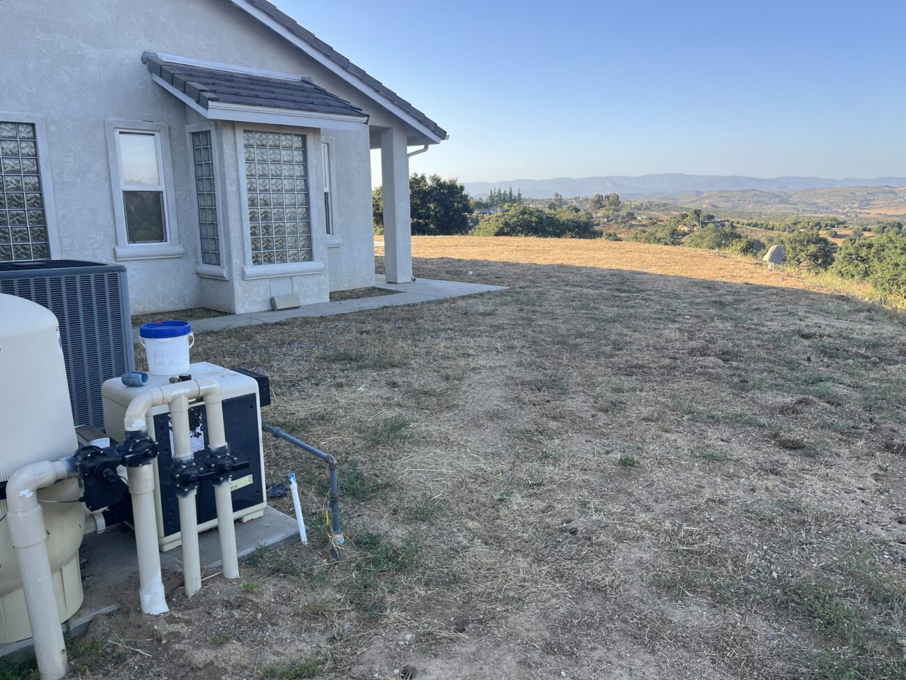 Residential rear yard showing a house exterior with a small concrete patio and adjacent area of dry dirt and sparse...